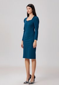 Robe ajustée teal avec un décolleté carré et des manches longues, présentant une silhouette cintrée. Des talons hauts à motifs étincelants complètent le look.