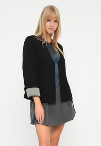Jonge vrouw met lang blond haar in een zwarte jasje met knopen, een blauwe top en een hoogwaist grijze geplooide rok tegen een witte achtergrond.