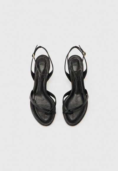 Μαύρες δερμάτινες σανδάλια με ανοιχτά δάχτυλα, λεπτά λουράκια που διασταυρώνονται και ρυθμιζόμενες αγκράφες στον αστράγαλο, εμφανιζόμενες από πάνω σε φωτεινό φόντο.