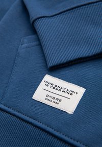 Tissu de sweat-shirt bleu montrant une étiquette blanche cousue avec le texte : "Votre seule limite est votre esprit" et "OMBRE depuis 1985."