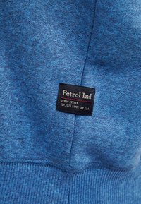 Sudadera azul con una tela texturizada y jaspeada. Presenta una etiqueta negra con texto blanco y rojo: "Petrol Ind" y "DENIM DRIVEN."