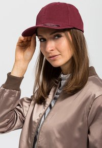 Tumeda baseball müts, millel on kumer ääreosa. Kanna isik sirgete pruunide juustega, hoides mütsi ääreosa.