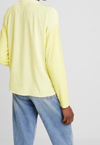 Person iført let gul langærmet top og løse lyseblå denimjeans, stående mod en ensfarvet grå baggrund, med ryggen mod kameraet.