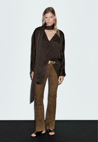 Top lungo in satin marrone a maniche lunghe con scollatura a V profonda e dettaglio a sciarpa, abbinato a pantaloni flare in suede marrone e sandali neri a scivolo.
