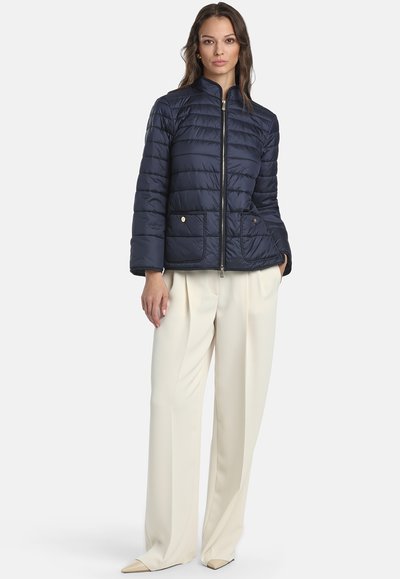 Donna che indossa una giacca trapuntata blu navy con zip frontale e pantaloni larghi crema, in piedi con una mano in tasca contro uno sfondo bianco.
