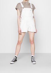 Persoon die witte denim overallshorts met rafelige zomen draagt, een beige t-shirt, witte sokken en zwarte hoge sneakers, staand op een grijze vloer.