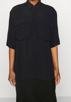 Camicia nera con colletto button-down, maniche corte e due grandi tasche anteriori. Il tessuto ha una texture liscia ed è caratterizzato da una vestibilità ampia.