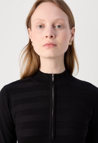 Černý pletený top s vysokým límcem a zipovým zapínáním. Má vodorovný pruhovaný vzor. Modelka nosí malé zlaté obdélníkové náušnice.
