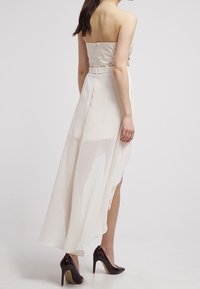 Olkaimeton, vaaleankermanvärinen iltapuku, jossa on tiukka vartalo-osa, läpinäkyvä päällyskangas, korkea-matala helma ja monimutkainen kukka-ompelu selkäpuolella.