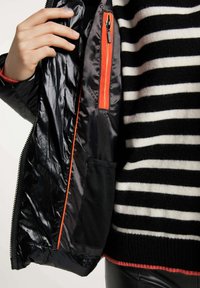Sort puffet jakke med blank tekstur, der har en indvendig lomme med en orange lynlås og en mesh-lomme. Båret over en sort og hvid stribet sweater.