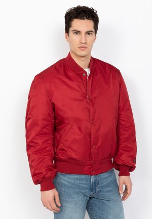 Red nylon bomber jacket features ribbed collar and cuffs, front snap closure, and side pockets. Worn over a white shirt with blue jeans.