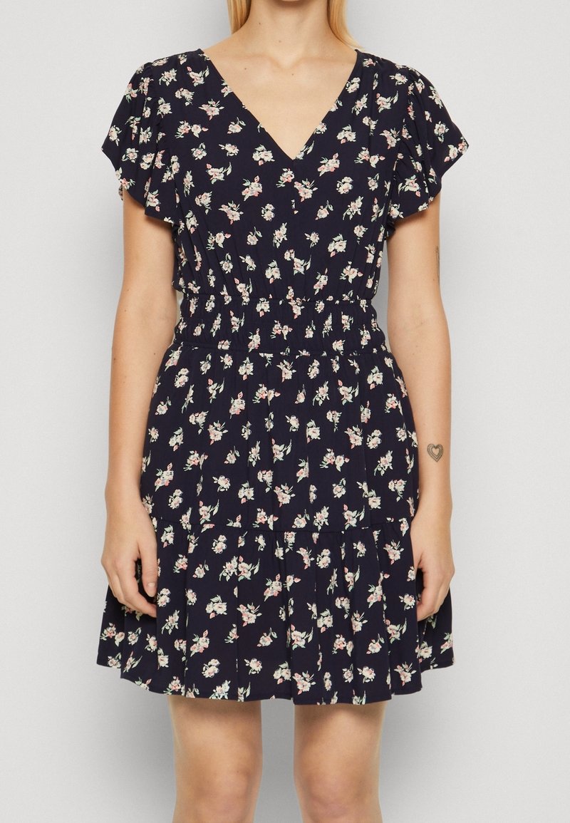 Femme portant une robe florale bleu marine à manches courtes, avec un décolleté en V et une taille élastique, debout devant un fond clair uni.