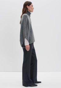 Frau, die im Profil steht und einen grauen hochgeschnittenen Poncho über weißen langen Ärmeln, schwarzen weit geschnittenen Hosen und schwarzen spitzen Schuhen trägt.