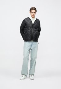 Jauns vīrietis melnā pārklāta jaka, baltā rāvējsl�ēdzēja džemperī, gaiši zilās plaša stila džinsās un baltās sporta kurpēs, stāv pret vienkāršu fonu.