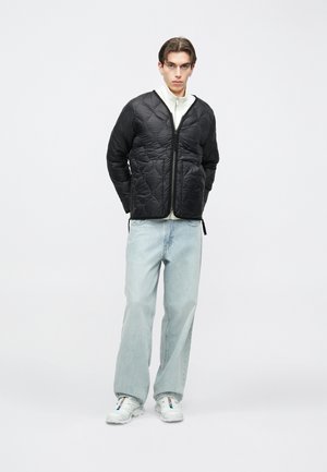 Noor mees, kes kannab musta tikitud jopet, valget tõmblukuga ülakeha, hele siniseid laiade säärega teksapükse ja valgeid tossusid, seisab ühevärvilise tausta ees.