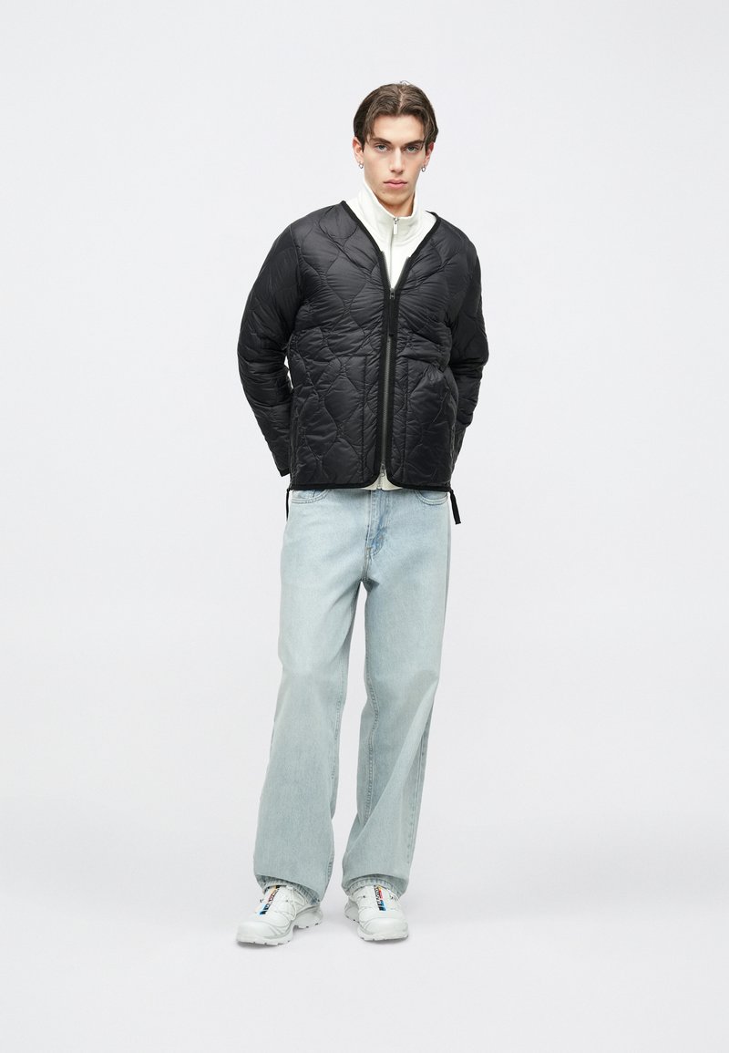 Jauns vīrietis melnā pārklāta jaka, baltā rāvējslēdzēja džemperī, gaiši zilās plaša stila džinsās un baltās sporta kurpēs, stāv pret vienkāršu fonu.