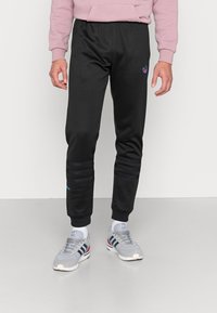 Svarta sweatpants med elastisk midja, texturerade ränder på knäna och en liten blå logodetalj. Kombinerat med grå atletiska skor.