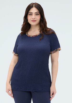 Kobieta o długich brązowych włosach, ubrana w granatowy, teksturowany top z krótkim rękawem z wielokolorową, koralikową lamówką na kołnierzu i rękawach, stojąca na jasnym tle.