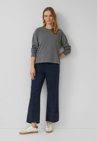 Femme se tenant contre un mur gris, portant un pull gris, un pantalon large bleu marine et des baskets blanches avec des semelles en caoutchouc.