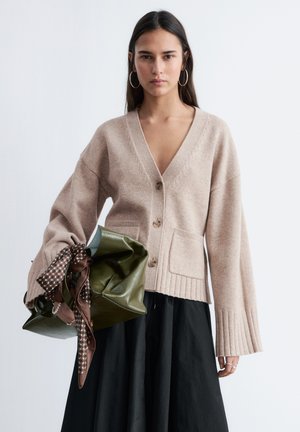 Woman wearing beige buttoned cardigan and black skirt, holding large green handbag decorated with brown patterned scarf.