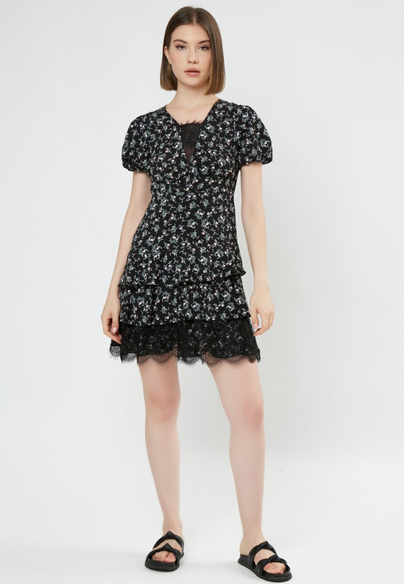 Vestido negro con flores que presenta mangas abullonadas, acentos de encaje, volantes en capas y un escote en V. Combinado con sandalias planas negras.