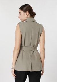 Γυναίκα με σκούρα μαλλιά σε χαμηλό κότσο, φοράει αμάνικο μπεζ γιλέκο με ζώνη και μαύρο παντελόνι, γυρισμένη πλάτη σε απλό φόντο.