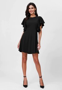 Robe noire courte avec des manches volantes, corsage ajusté et jupe évasée. Fabriquée en tissu lisse, associée à des talons hauts noirs.