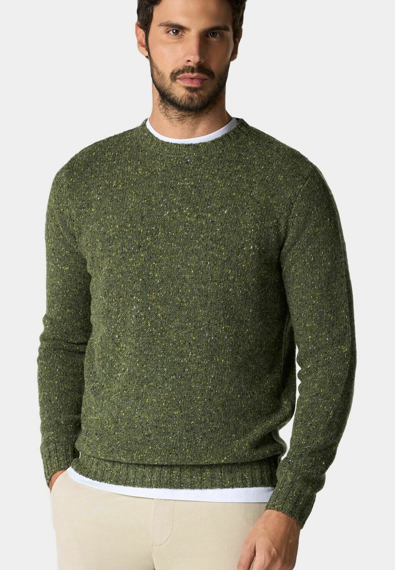 Dunkelgrüner Strickpullover mit strukturierter Oberfläche und gelben Sprenkeln. Mit Rippbündchen und Saum, Rundhalsausschnitt und über einem weißen Hemd getragen.