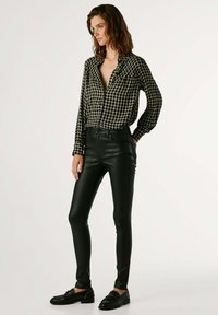 Camisa a cuadros negra y verde con diseño de botones, combinada con pantalones ajustados de cuero sintético negros y mocasines negros.