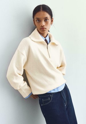 Pull en crème avec un col montant, fermeture éclair sur le devant et poignets côtelés, superposé sur une chemise bleu clair, associé à un jean sombre.