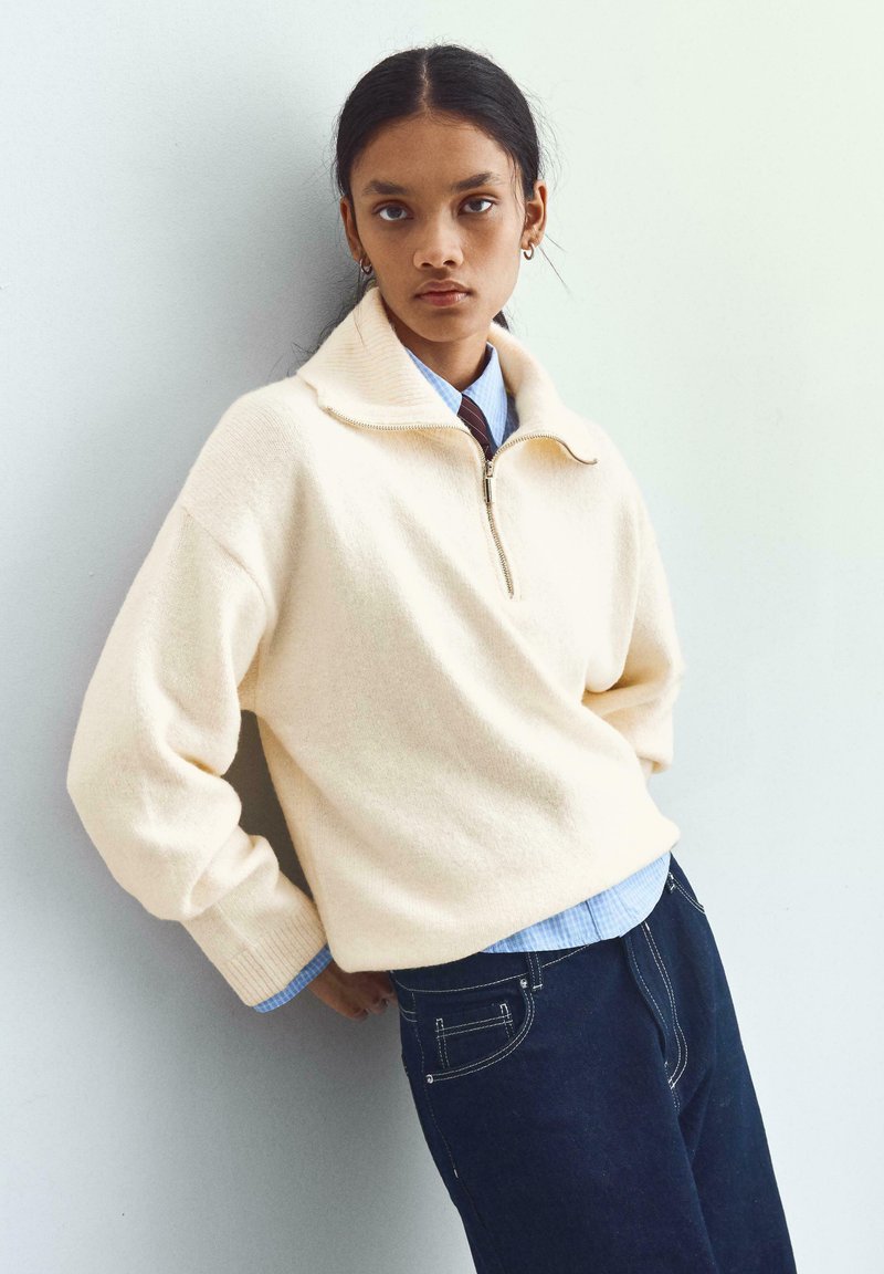 Krémový svetr s vysokým límcem, zipem na přední straně a žebrovanými manžetami, vrstvený přes světle modrou košili, doplněný o tmavé džíny.