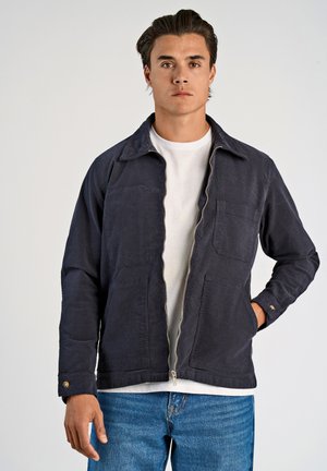 Giacca di tessuto blu navy con zip frontale, due tasche sul petto e polsini con bottone. Indossata sopra una t-shirt bianca e abbinata a jeans di denim.