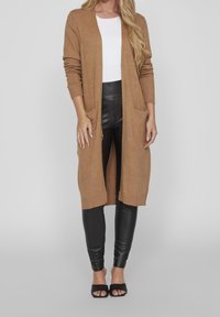 Cardigan lungo color cammello realizzato in materiale morbido, con una texture a coste, apertura frontale e due tasche anteriori. Abbinato a leggings neri.