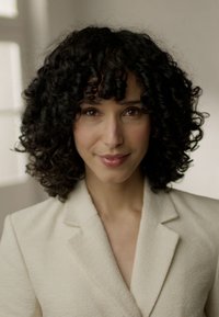 Kremowa marynarka z kołnierzem, wykonana z teksturowanej tkaniny, z głębokim dekoltem w kształcie litery V i bujnymi, kręconymi włosami. Miękkie oświetlenie podkreśla naturalne cechy.