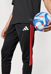 Μαύρο αθλητικό παντελόνι με κόκκινες ρίγες, με λευκό λογότυπο Adidas. Κρατώντας πολύχρωμη ποδοσφαιρική μπάλα με μπλε, κόκκινα και λευκά σχέδια.