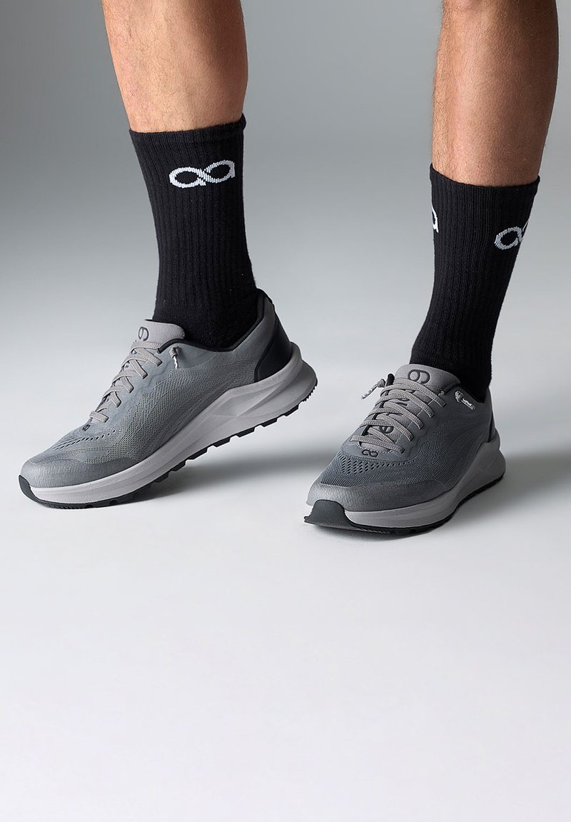 Baskets de sport grises en tissu texturé, avec des accents noirs et une semelle blanche. Associées à des chaussettes côtelées noires arborant un symbole d'infini blanc.