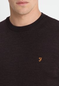 Maglione marrone scuro con collo rotondo, realizzato in tessuto lavorato a maglia. Presenta un piccolo logo arancione sul petto. La trama appare liscia e lucida.
