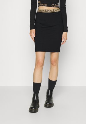Personne portant une jupe noire, un crop top noir à manches longues avec des bandes visibles du logo Calvin Klein Jeans, des chaussettes noires et des bottines noires.