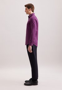 Lila Langarmhemd mit Button-Down-Kragen, kombiniert mit schwarzen maßgeschneiderten Hosen und schwarzen Schuhen, von der Seite betrachtet.