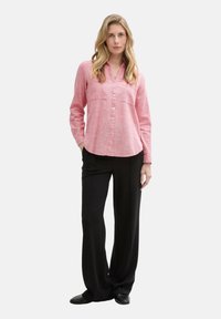 Donna in piedi che indossa una camicia rosa a maniche lunghe con bottoni e pantaloni neri larghi, abbinati a scarpe nere, su uno sfondo neutro.