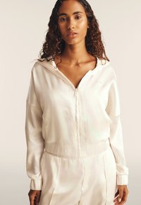 Felpa con zip color crema in satin, dotata di cappuccio, vita e polsini elasticizzati. Presenta una leggera lucentezza e un logo ricamato sul petto.