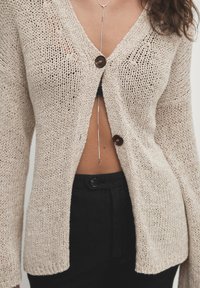 Beige gestrickte Strickjacke mit V-Ausschnitt, ausgestattet mit einer braunen Knopfverschluss und lockeren Ärmeln. Die Textur ist offen gestrickt und lässt eine darunterliegende Schicht hervorschauen.