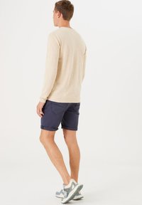 Beige langärmliges Hemd mit rundem Halsausschnitt, kombiniert mit marineblauen hochgekrempelten Shorts und hellen Sneakern mit türkisfarbenen Akzenten.