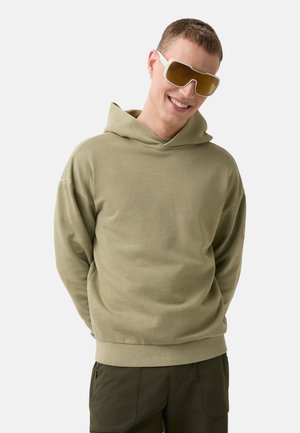 Kapuzensweatshirt in hellem Olivgrün, aus weichem Stoff mit lockerer Passform und gerippten Säumen. Getragen mit großen, hellen Sonnenbrillen.