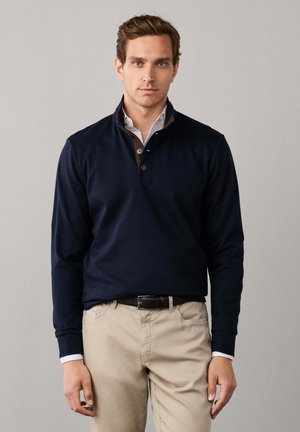 Man wearing navy blue pullover over white shirt and beige pants standing against plain light background.