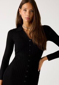 Abito nero a coste con maniche lunghe, colletto polo e chiusura a bottoni frontale. Presenta una vestibilità slim e una texture morbida.