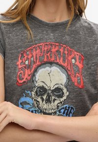 Camiseta de algodón gris con un gráfico rojo que lleva el texto "Superdry" sobre un detallado diseño de calavera negra y llamas azules. Mangas cortas, escote redondo.
