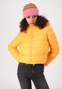 Doudoune jaune au design court avec des coutures diagonales, assortie à un bonnet en tricot rayé rose et marron.