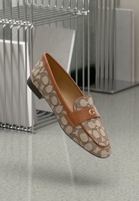 Brown patterned loafers with a tan leather strap, featuring a gold logo accent and a low black heel against a minimalist background.