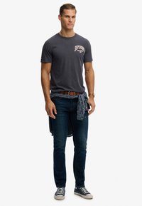 Camiseta de manga corta gris oscuro con el logo "Superdry", combinada con jeans azules medios. Una camisa a cuadros azul atada a la cintura. Zapatillas casuales.